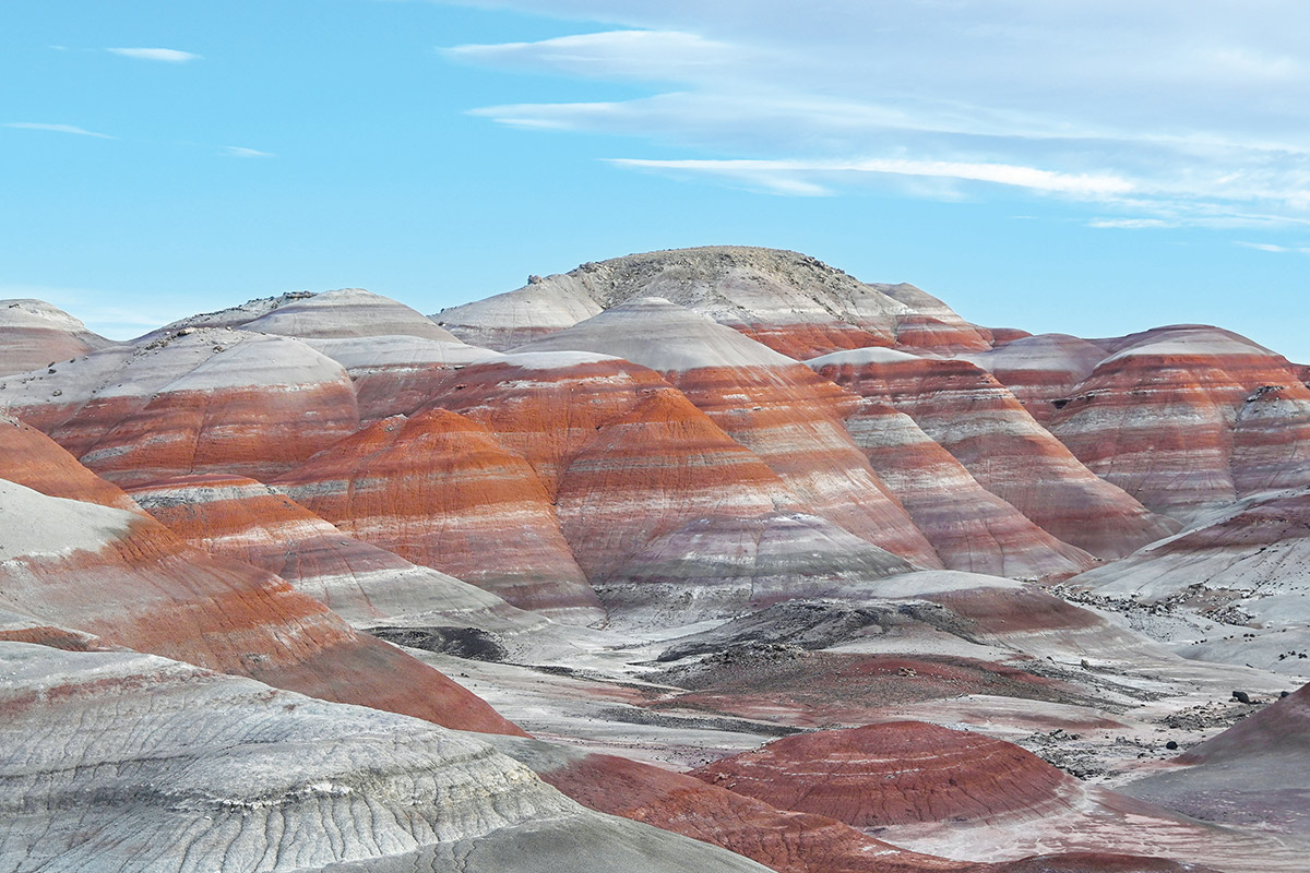 View to the north of variegated slopes of the Brushy Basin Member of the Jurassic-age Morrison Formation in the Bentonite Hills. Striped orange, red, and gray hills under a pale blue sky.