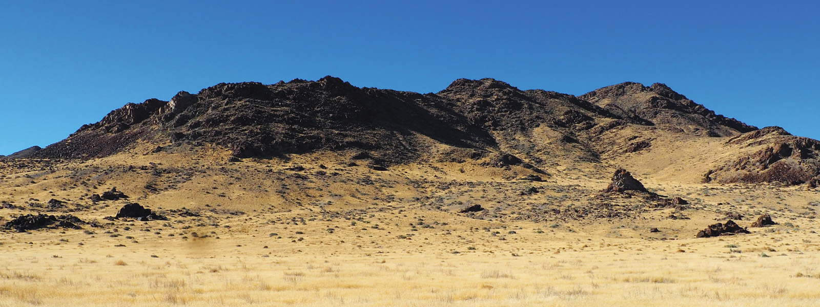 Approximately 18,000 years ago, waves etched the Bonneville Shoreline near the base of Desert Mountain. The shoreline is the flat area just below the dark outcrops of volcanic rock that erupted explosively when the caldera was formed. A mountain landscape topped with dark black rocks and dry grass in the foreground.